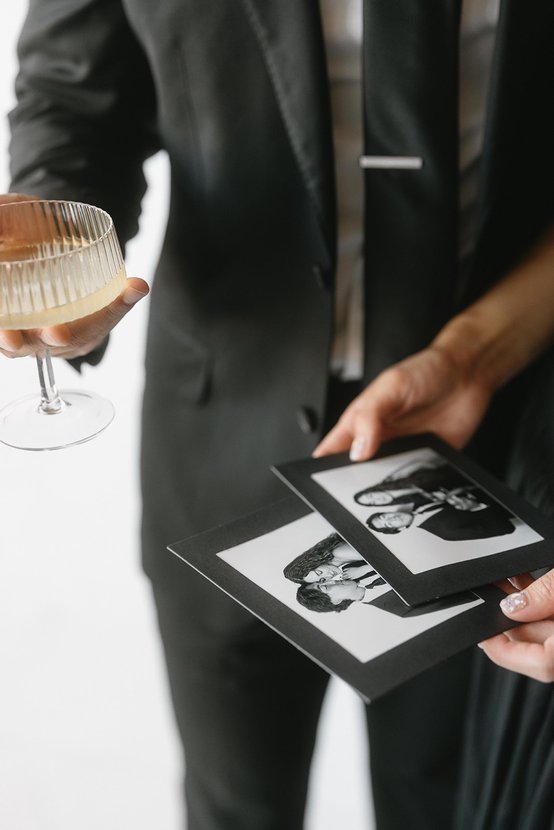 A person in a suit holding a coupe glass, another holding photo prints